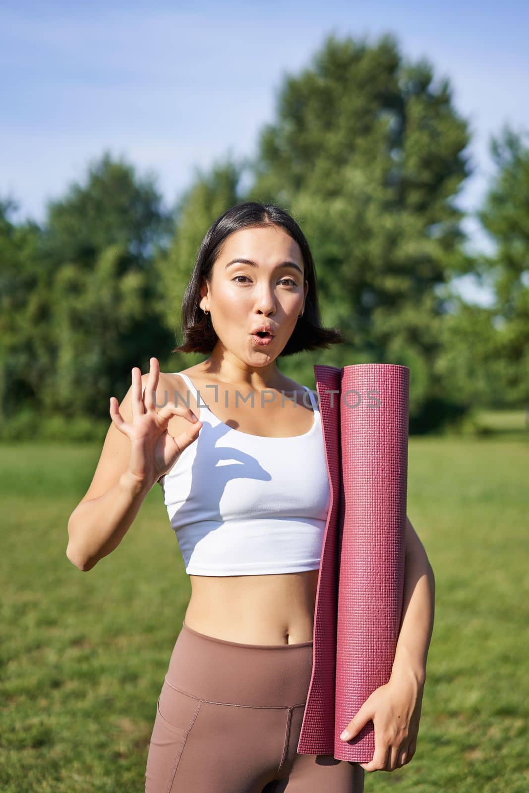 Excited young woman standing with sports mat, yoga clothes, shows okay sign, workout in park, wellbeing training session outdoors by Benzoix