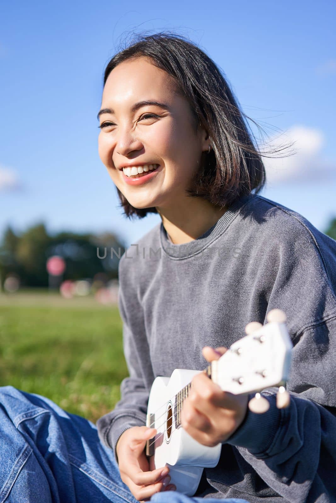 Vertical shot of happy korean girl sitting in park, learning how to play ukulele, singing and relaxing by Benzoix