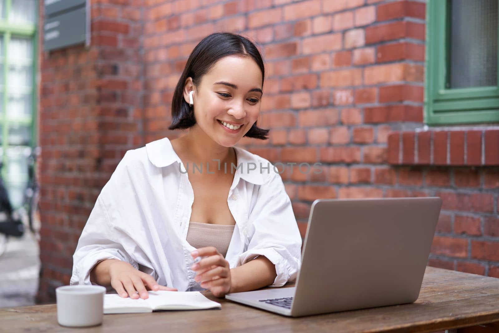 Young smiling asian woman with wireless earphones, sitting in coworking office space with laptop, doing homework, making notes.