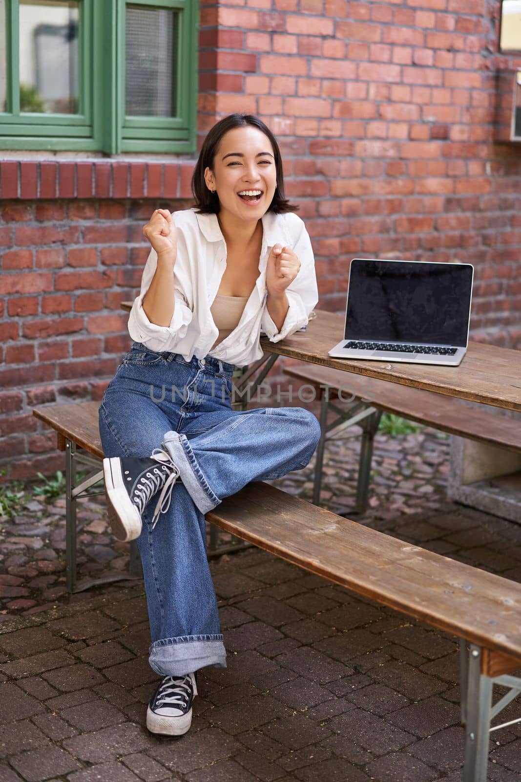 Vertical shot of enthusiastic asian girl, sits with laptop, screen is empty and blank, woman laughing and celebrating, triumphing by Benzoix