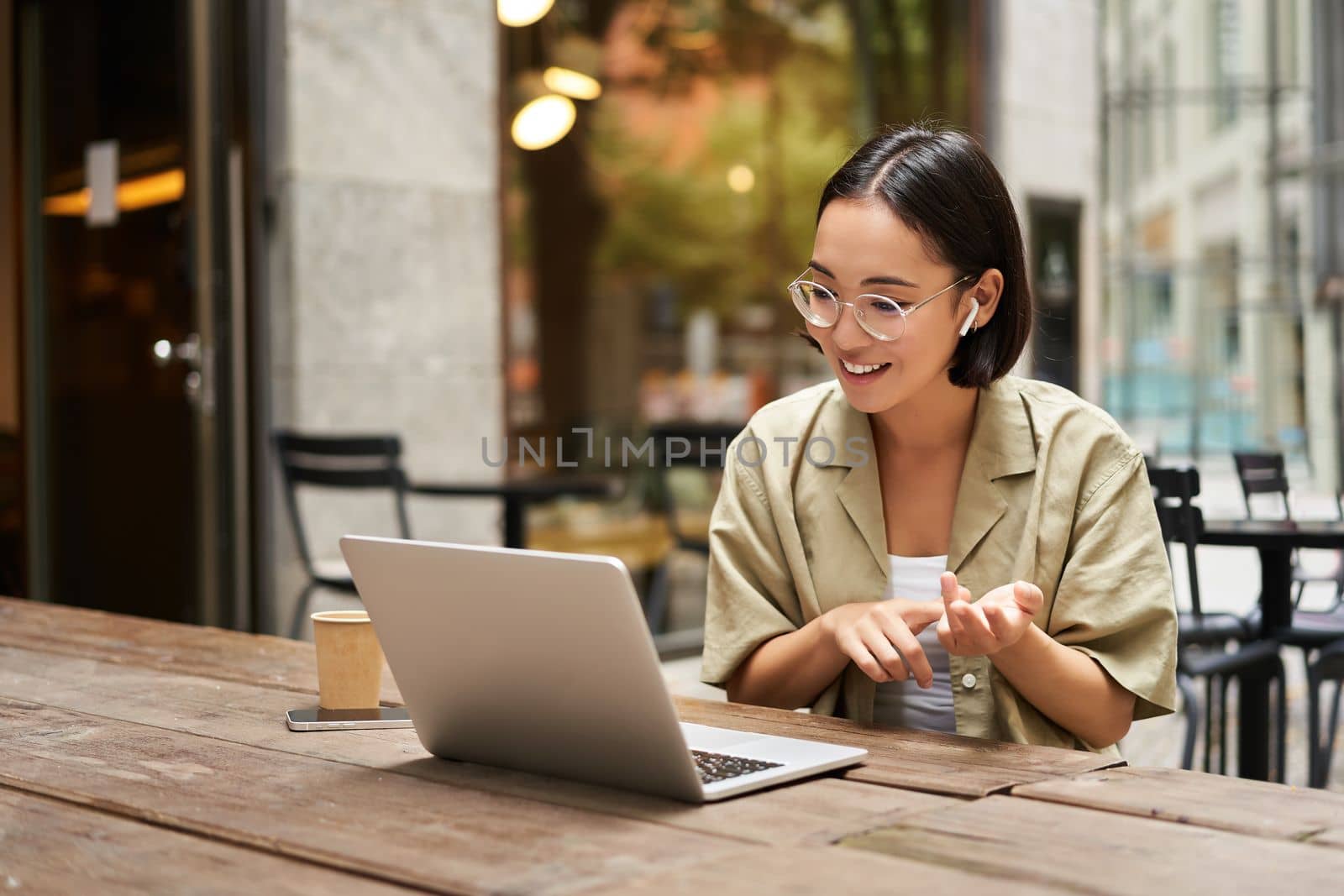 Young woman sitting on online meeting in outdoor cafe, talking to laptop camera, explaining something, drinking coffee. Digital nomad