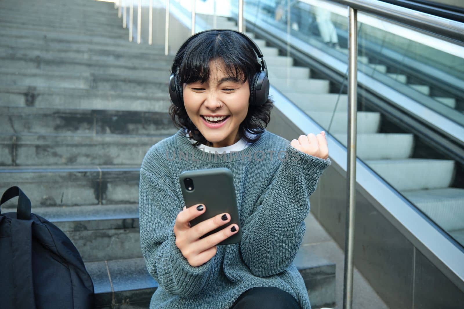 Enthusiastic korean girl in headphones, watches video and celebrates, looks at smartphone with triumphing, pleased face, say yes, sits on stairs in public place by Benzoix