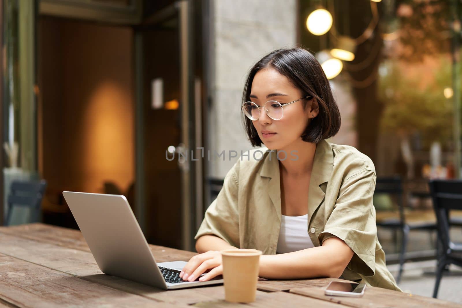 Young woman working in a cafe, using laptop and drinking coffee. Asian girl student with computer studying remotely, sitting on bench near shop by Benzoix