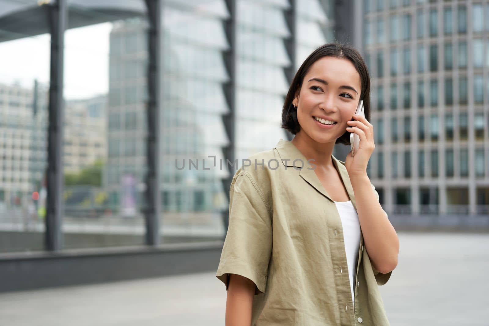 Portrait of asian girl smiling, talking on phone, making a call, standing on street near building and waiting for someone, answer telephone.