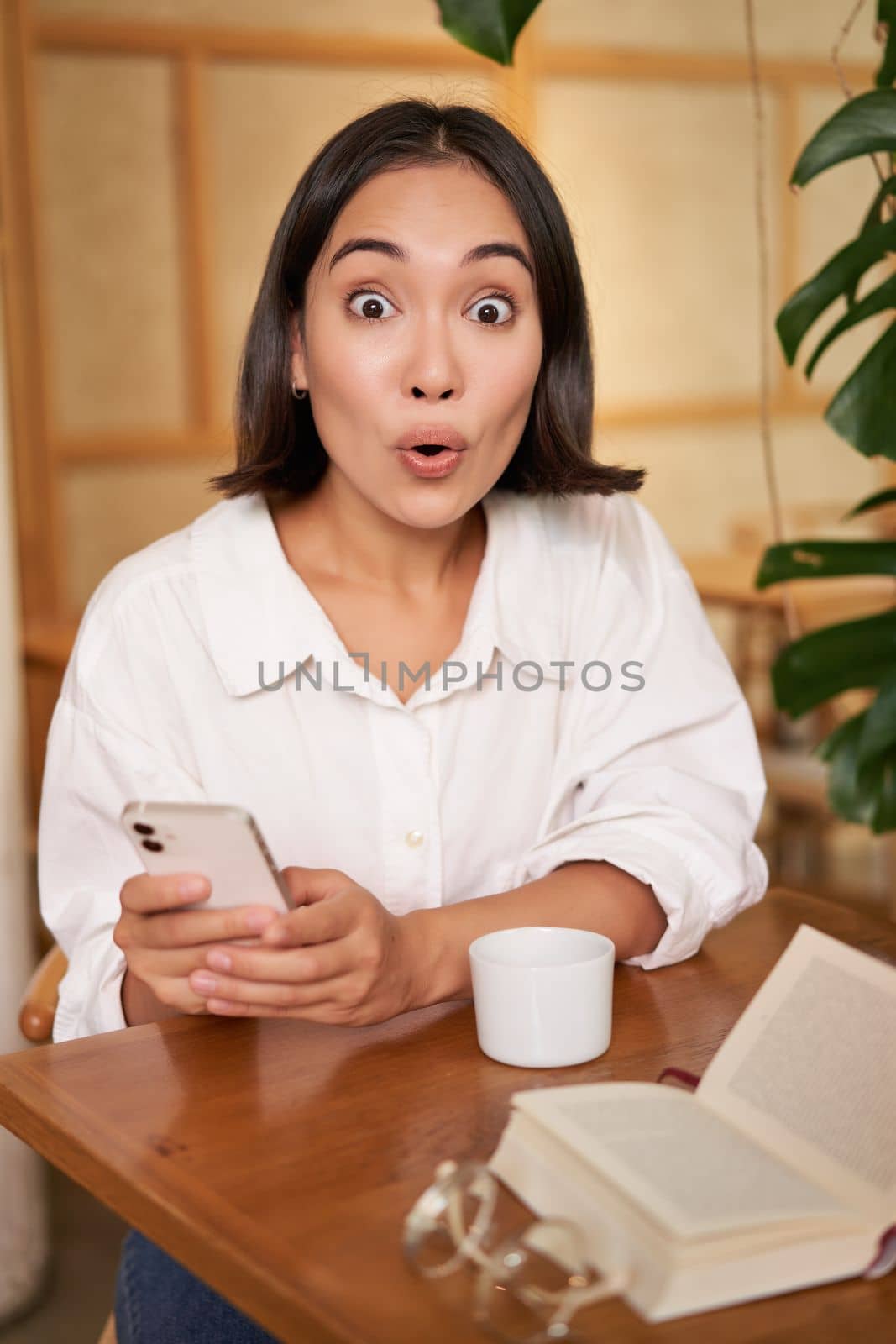 Portrait of asian girl looking surprised, holding mobile phone with amazed face, great news on smartphone, sitting in cafe with book by Benzoix