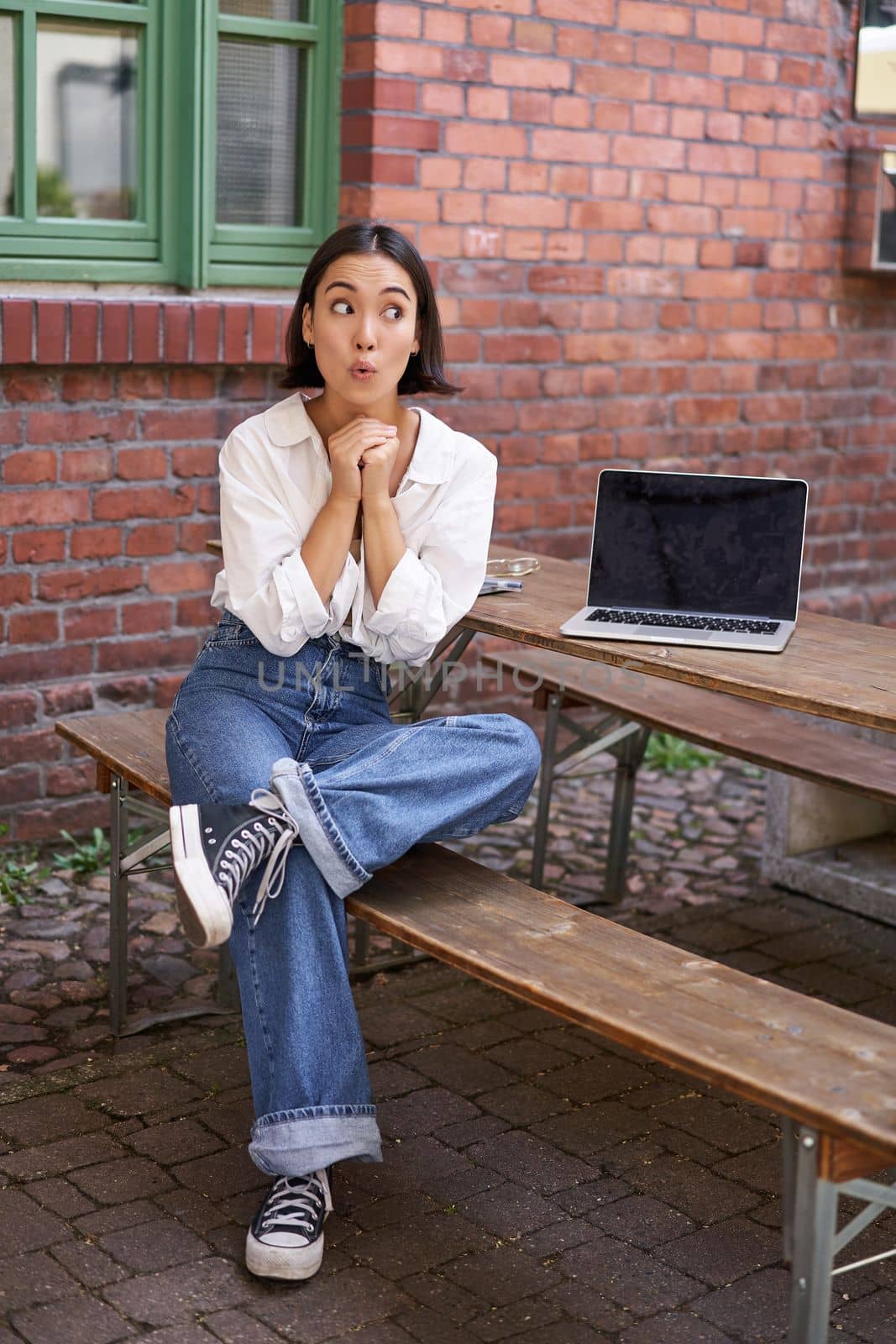 Full size portrait of asian girl sits with laptop, looks amazed and surprised and peeks at screen. Concept of people and urban lifestyle.