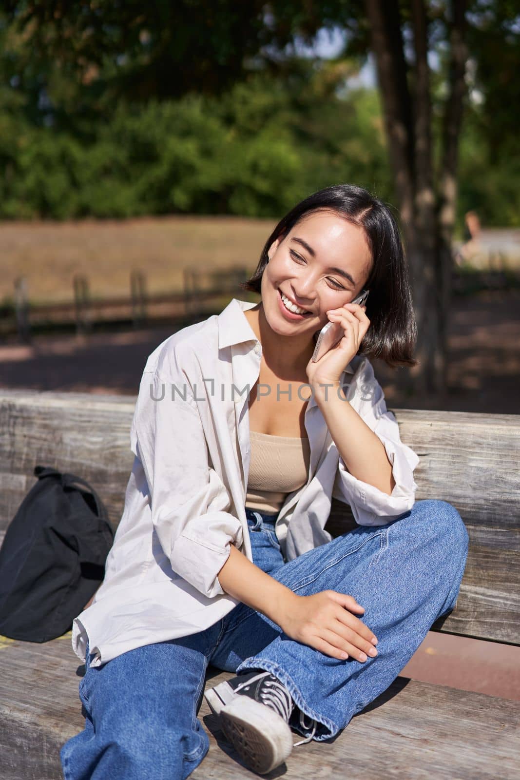 Smiling asian girl relaxing in park, talking on smartphone, having a mobile call while resting outdoors.