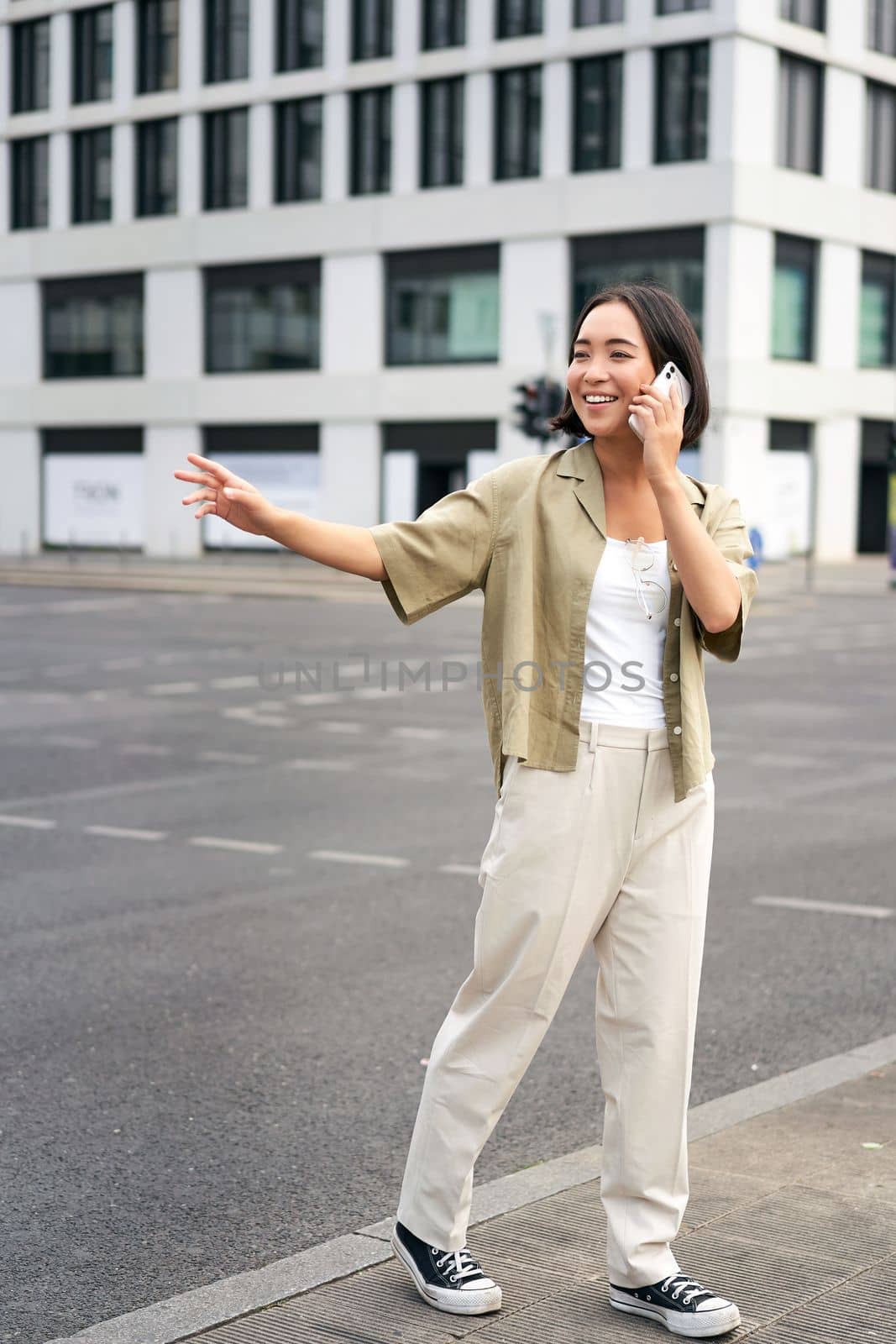 Young woman waving to taxi driver, calling or talking on mobile phone, order car ride, standing outdoors on street.