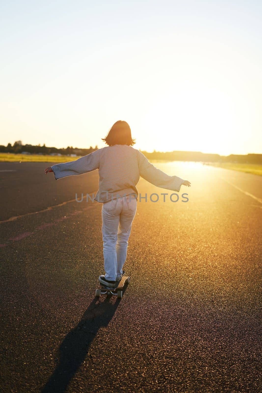 Rear view photo of young girl riding skateboard towards sunlight. Happy young woman on her cruiser, skating on longboard by Benzoix