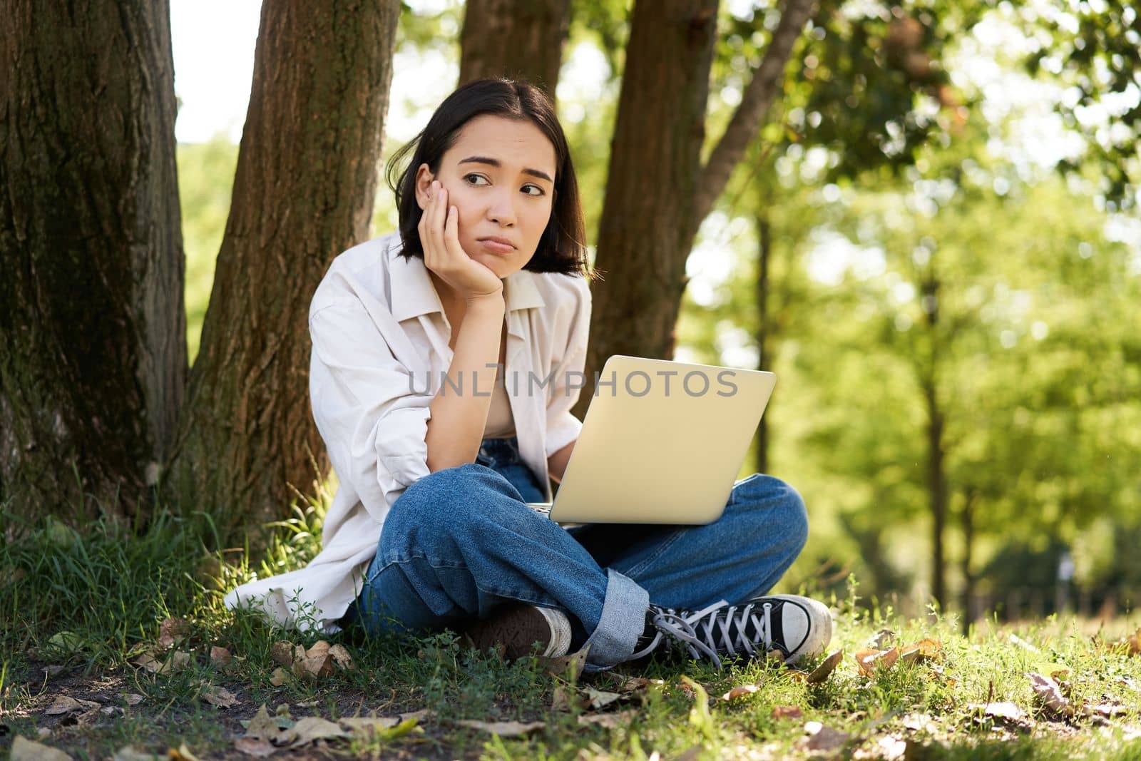 Portrait of young asian woman sitting in park near tree, working on laptop, using computer outdoors by Benzoix