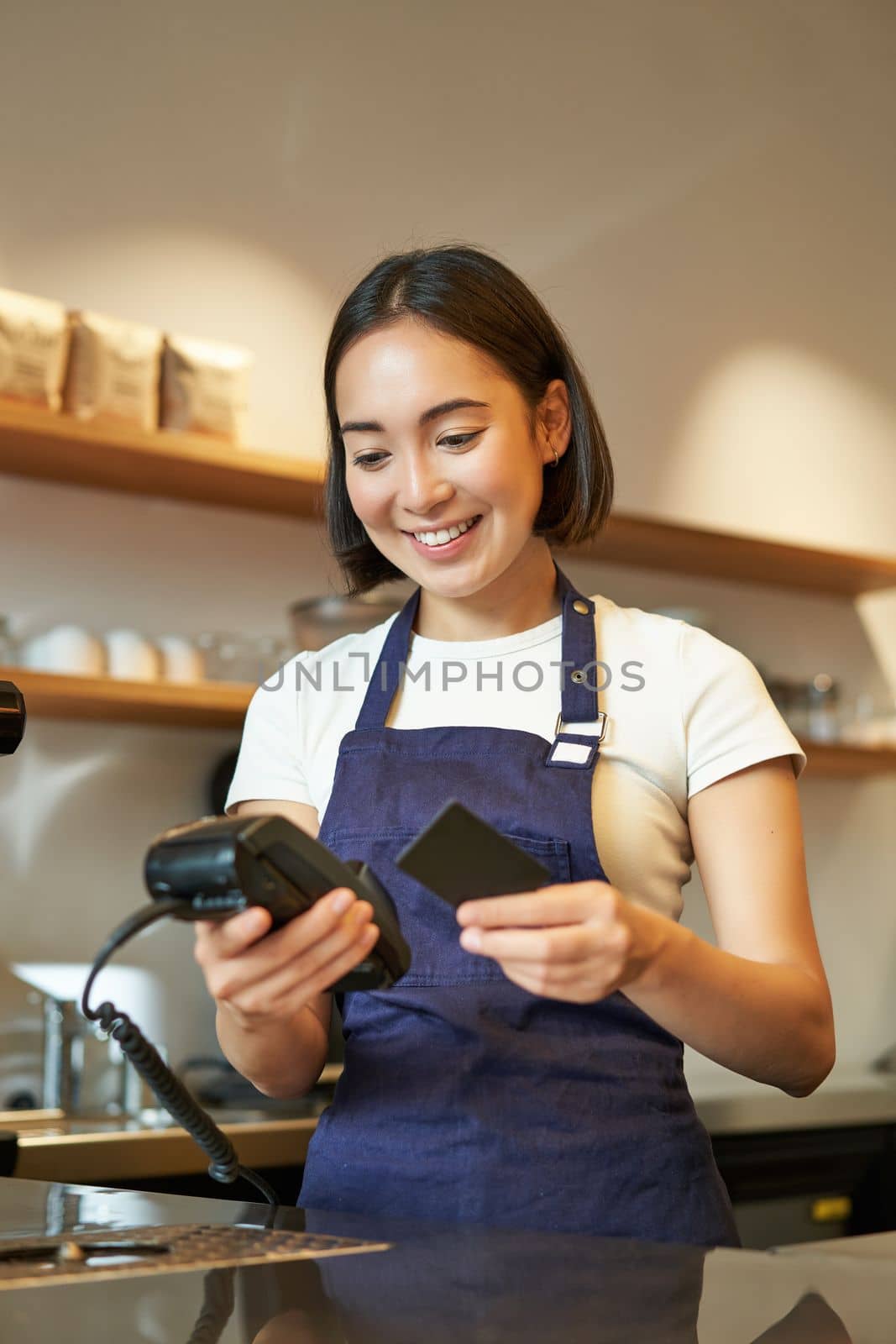 Portrait of smiling asian barista, coffee shop employee using POS terminal and credit card, helps client pay contactless in cafe by Benzoix