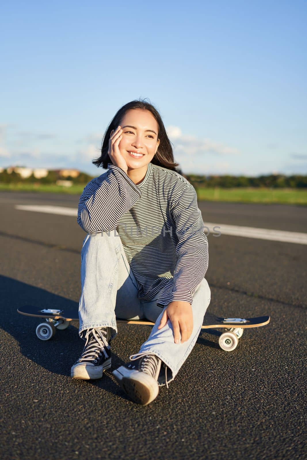 Vertical shot of asian girl skater, sits on her skateboard and smiles, enjoys sunny day, cruising on longboard on empty road outdoors.