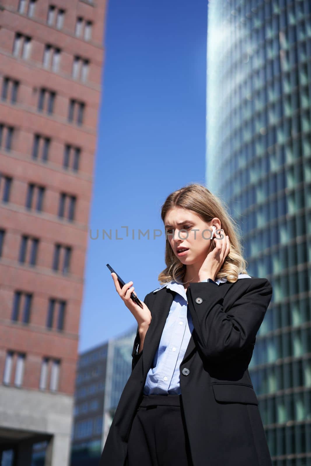 Businesswoman in wireless earphones looking at mobile phone, has concerned face expression, reads message on smartphone.