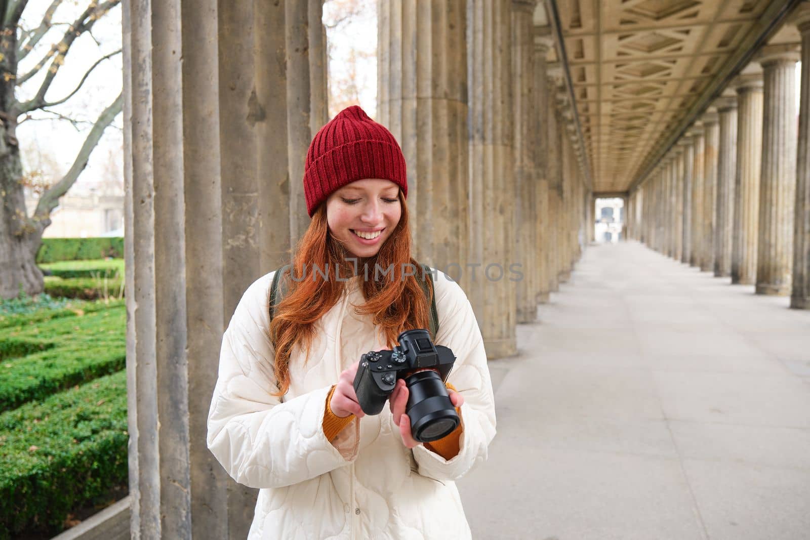 Young redhead female photographer, makes lifestyle shooting in city centre, takes photos and smiles, looks for perfect shot, makes picture by Benzoix