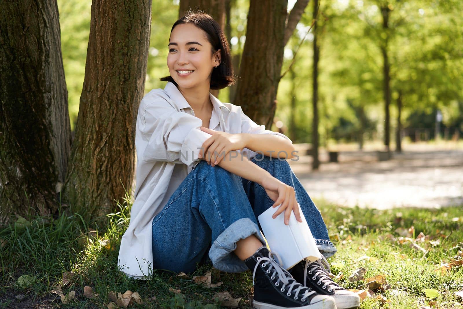 Woman sitting in park with her favourite book, leaning on tree under shade on sunny day, enjoying nature and calm relaxing atmosphere.