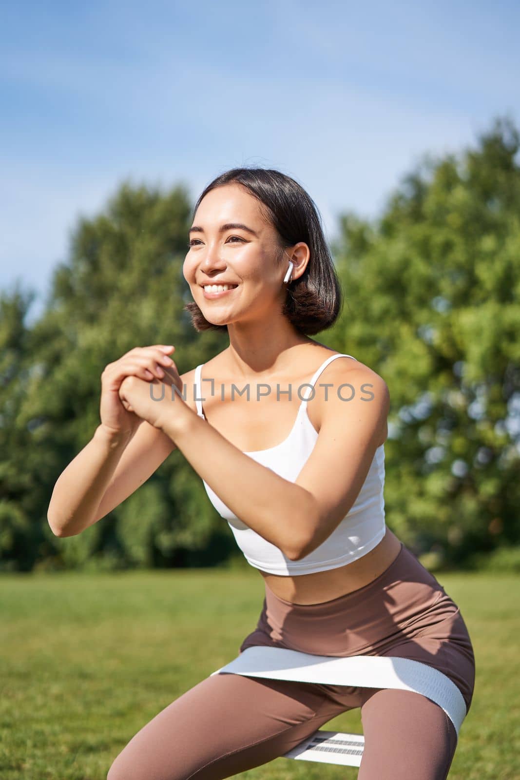 Asian fitness girl doing squats in park, using resistance band, stretching yoga rope for workout training on fresh air by Benzoix