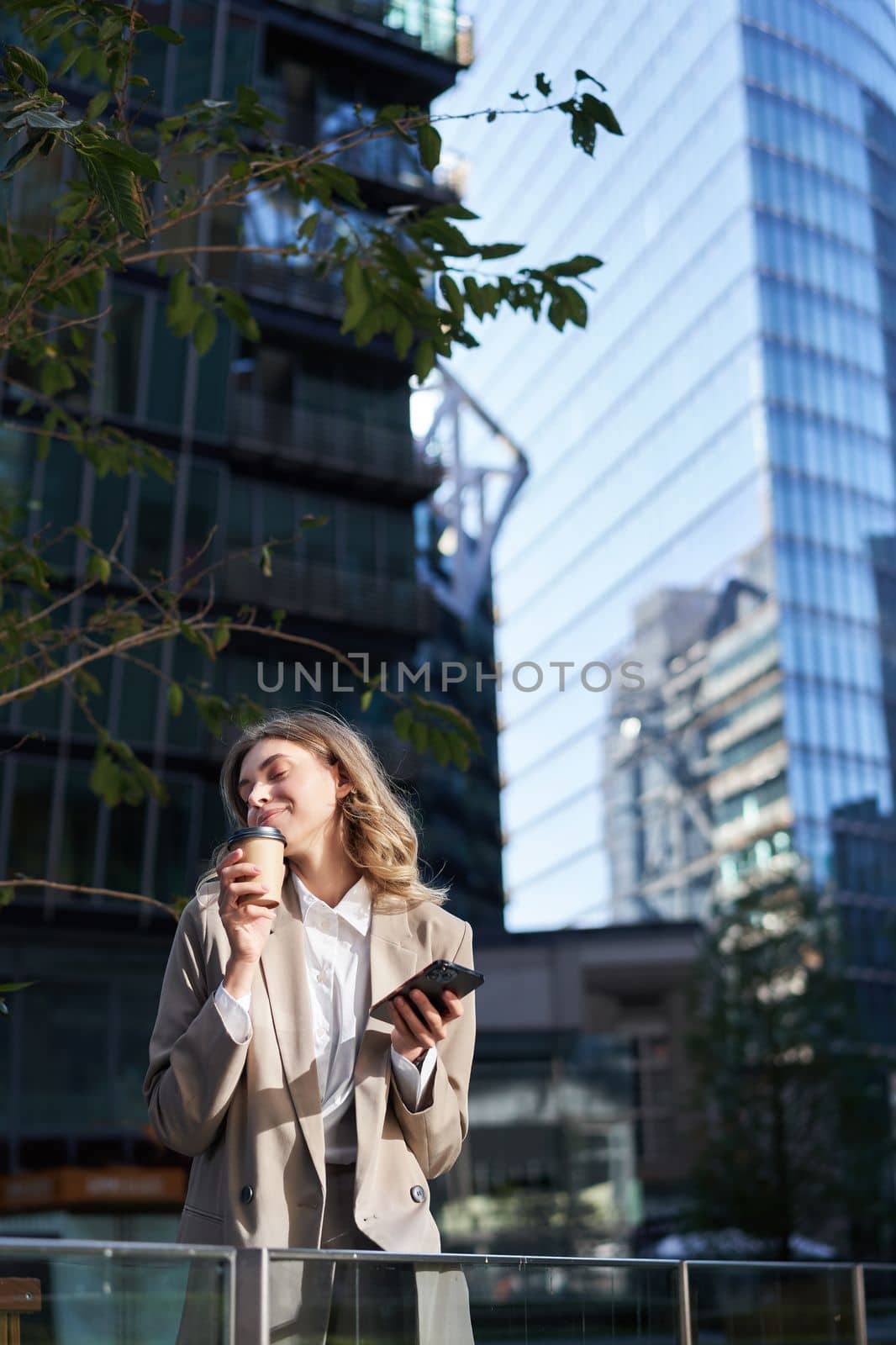 Enthusiastic businesswoman drinks her coffee takeaway on street, holds mobile phone, standing in beige suit, office outfit.