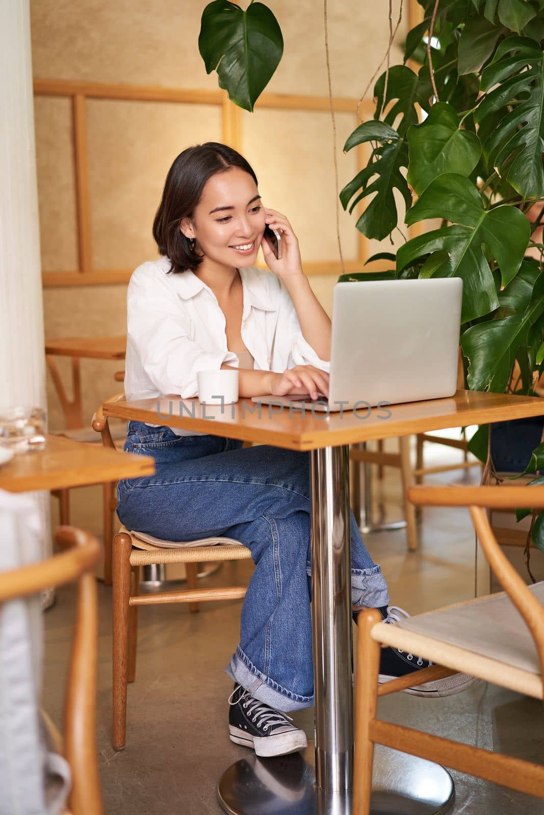 Stylish young woman answer phone call, sitting in cafe with laptop, working and taling on smartphone.