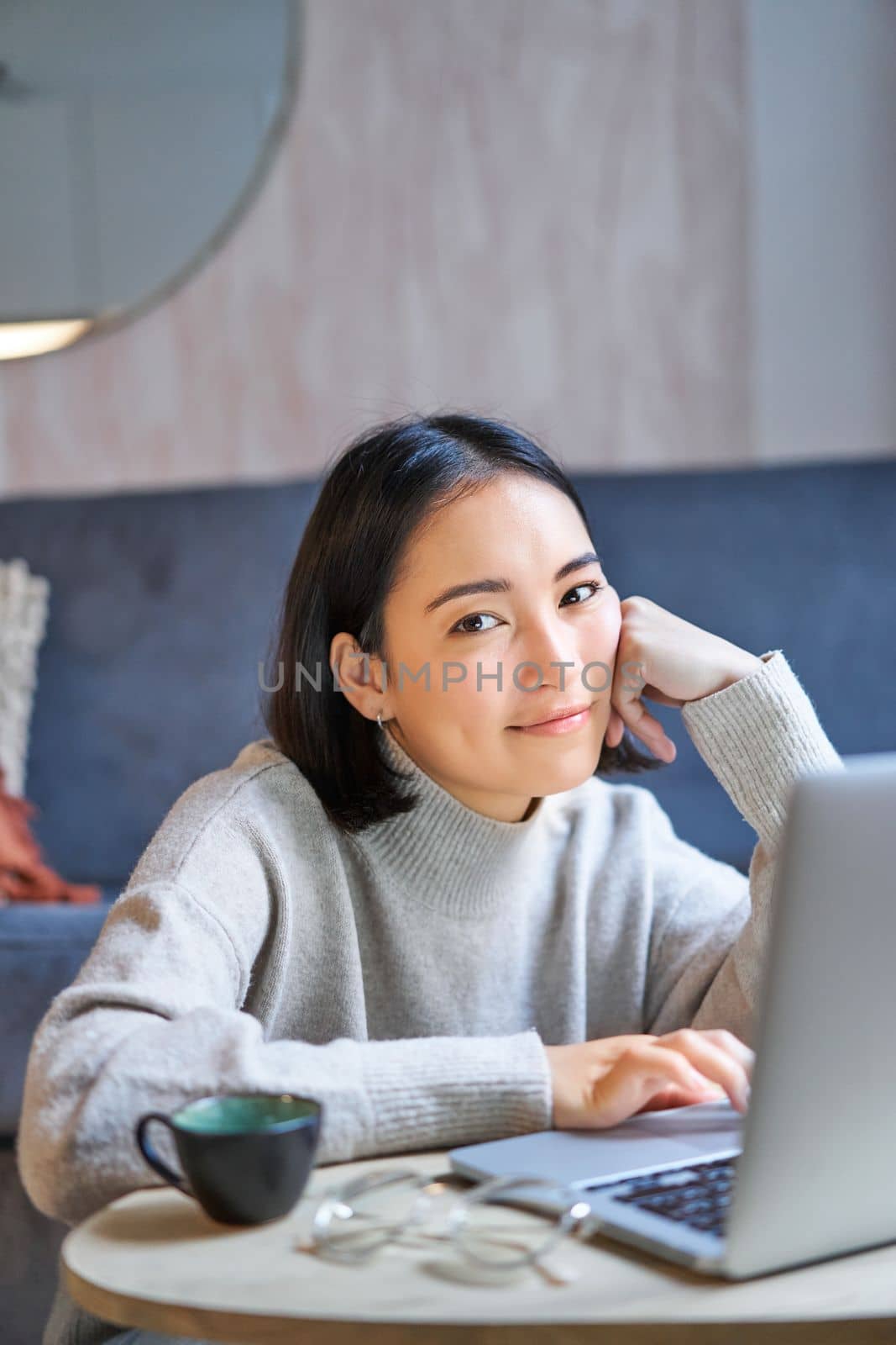 Vertical shot of young asian woman looking tired, smiling with exhausted expression, working from home on her laptop by Benzoix