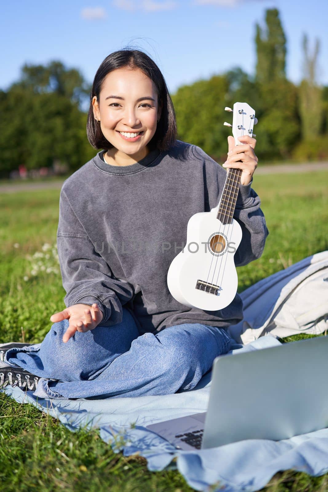 Vertical shot of happy asian girl plays instrument, shows her ukulele at laptop camera, video chats about music, teaches how to play, sits in park outdoors by Benzoix