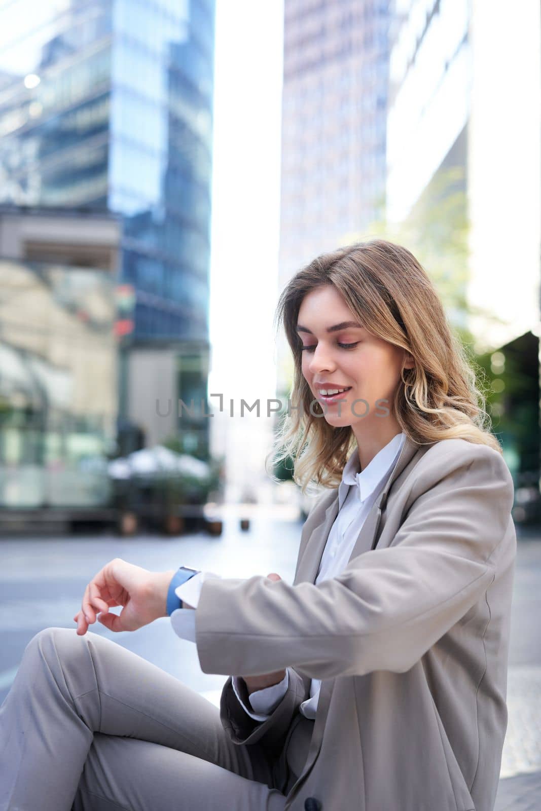 Vertical shot of smiling woman reading message on her digital watch, looking at wrist, checking time, wearing office suit by Benzoix