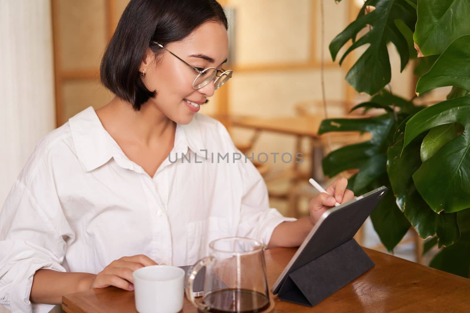 Portrait of beautiful young woman sitting in cafe, drinking coffee and drawing diagram on tablet with graphic pen, smiling pleased.