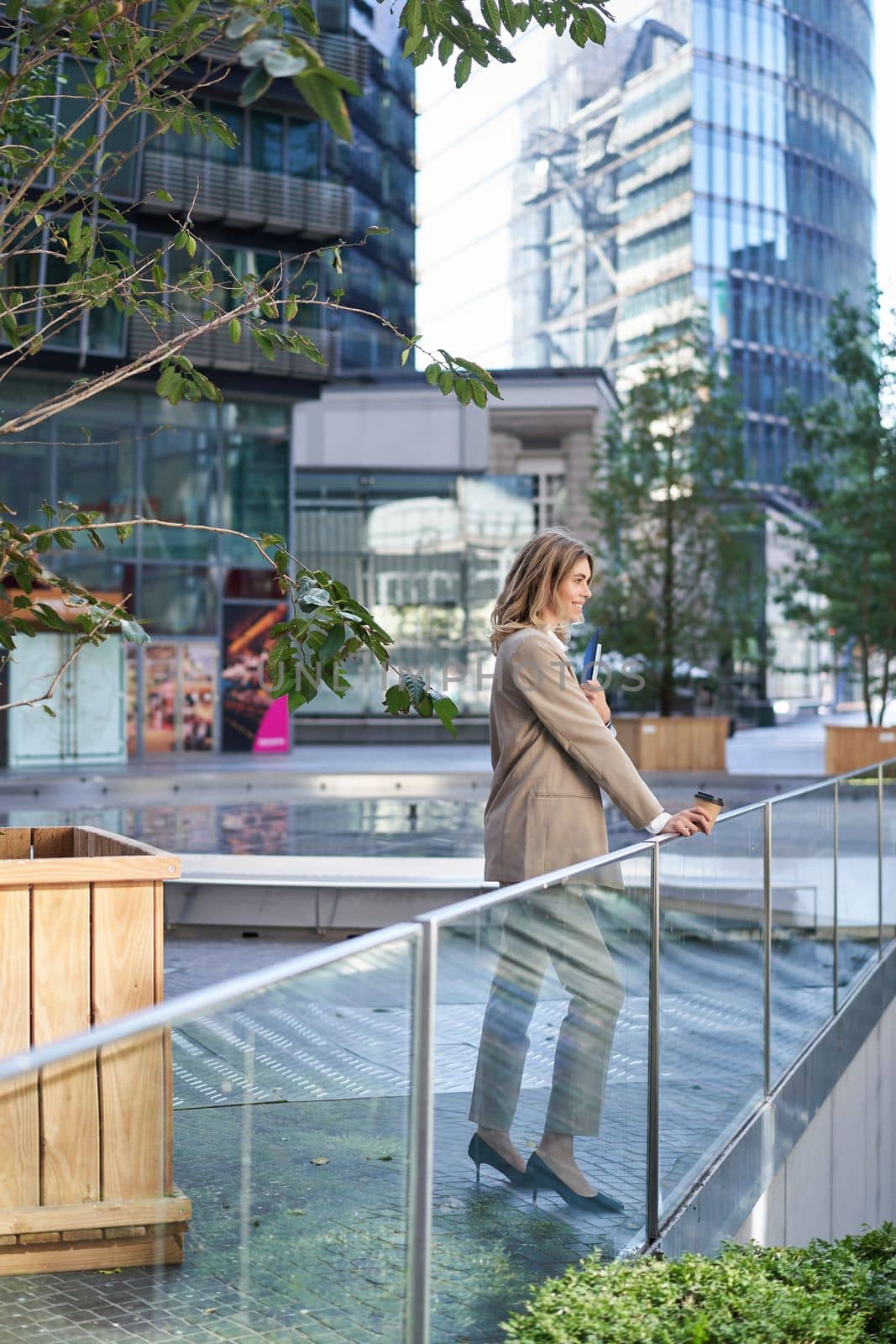 Vertical shot of corporate woman in beige suit standing outside on street, drinks morning coffee takeaway before work.