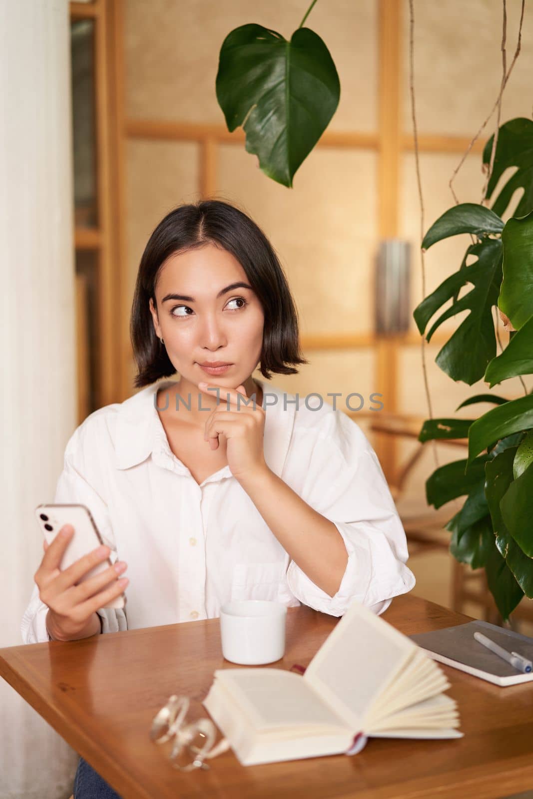 Beautiful young woman, 25 years old, thinking, holding smartphone and looking thoughtful, sitting in cafe, deciding smth.