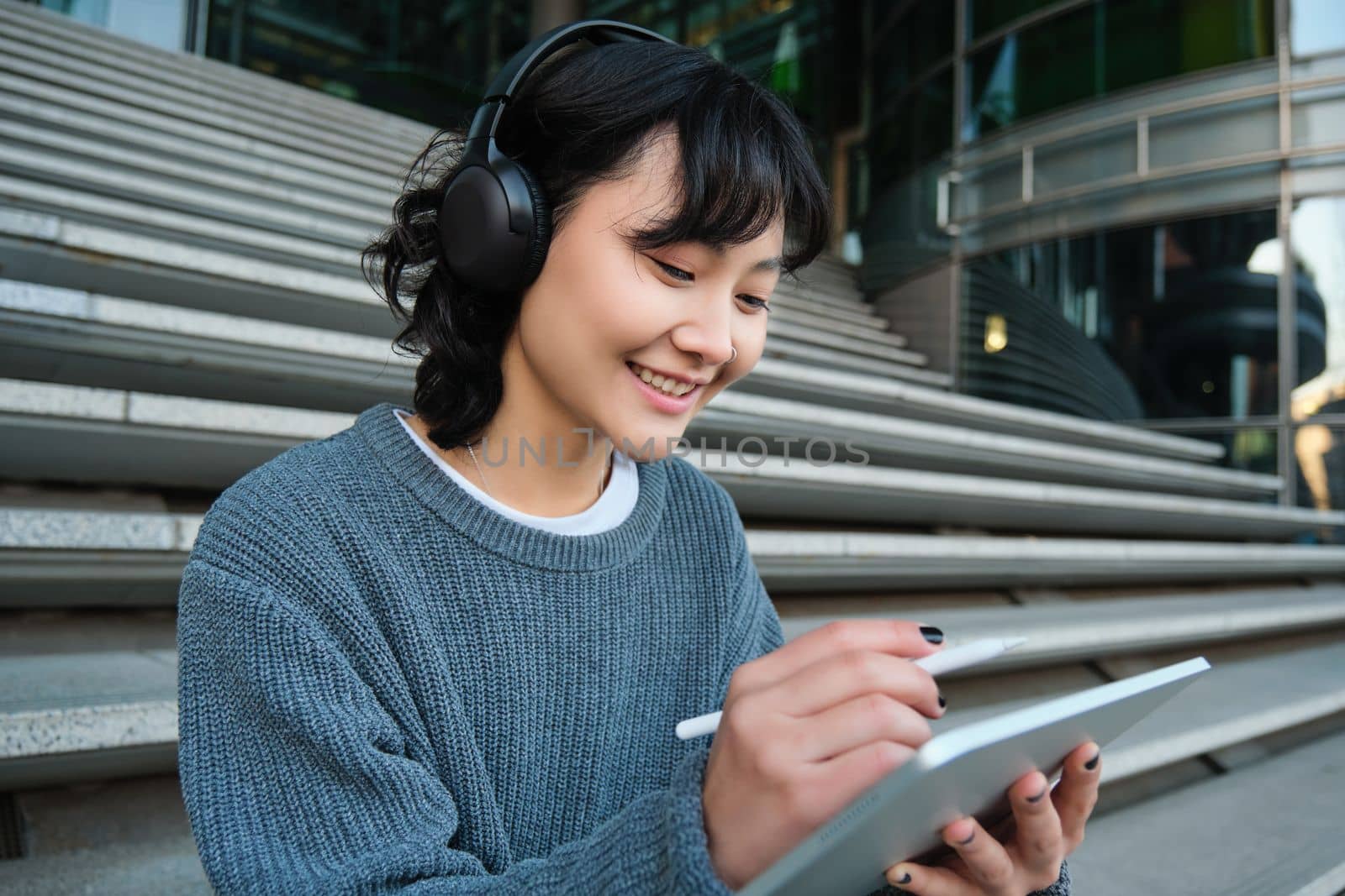 Young woman draws on her tablet, listens to music in headphones. Asian girl sits on stairs and does graphic design project, sits on street stairs.