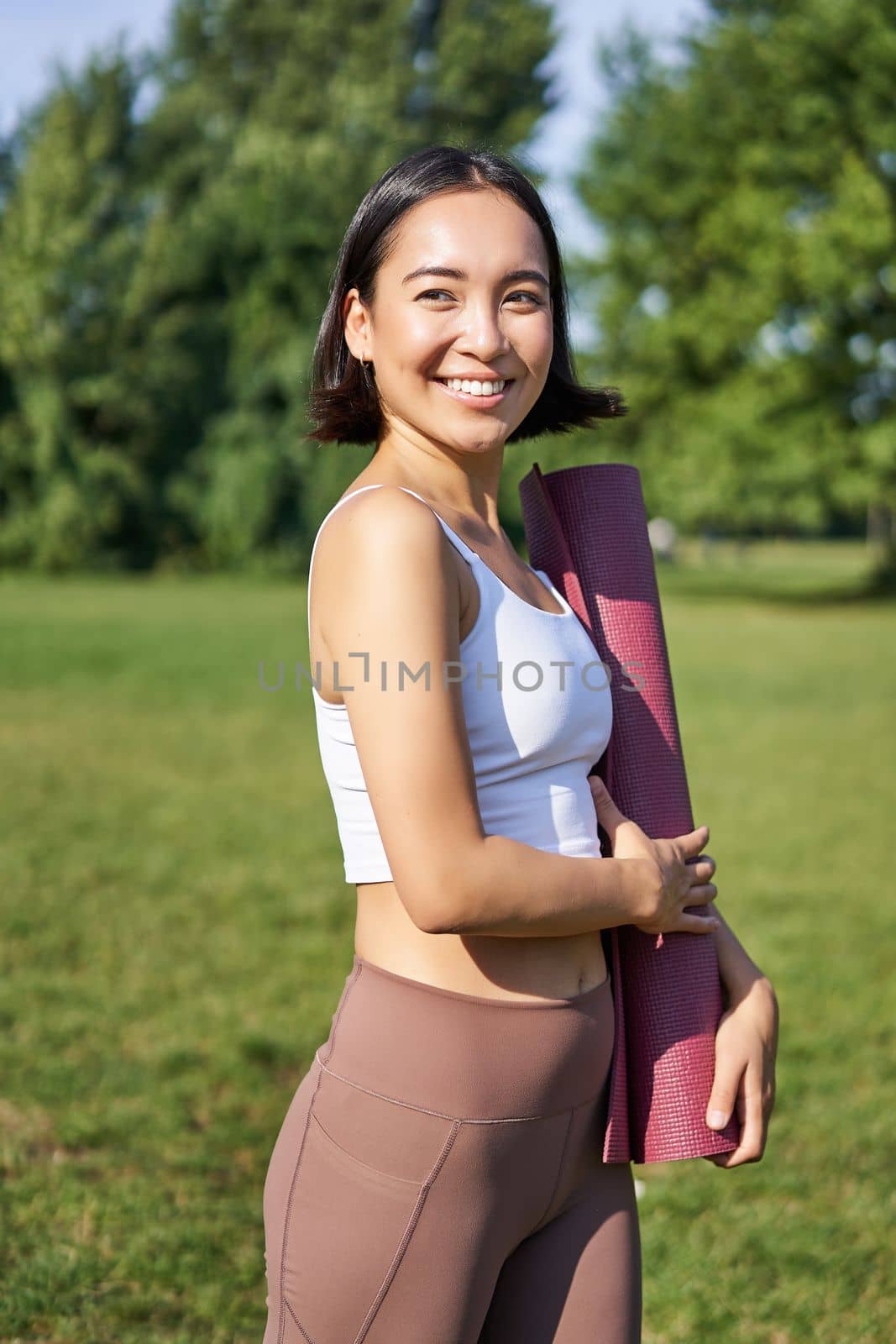 Smiling fitness girl with rubber mat, stands in park wearing uniform for workout and sport activities, does yoga outdoors on lawn by Benzoix