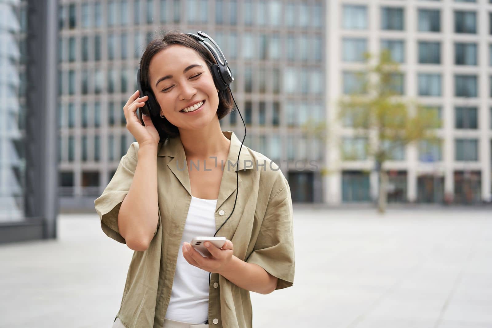 Streaming services concept. Happy asian girl listens music in headphones, holds mobile phone, choosing track or podcast, walking on street.