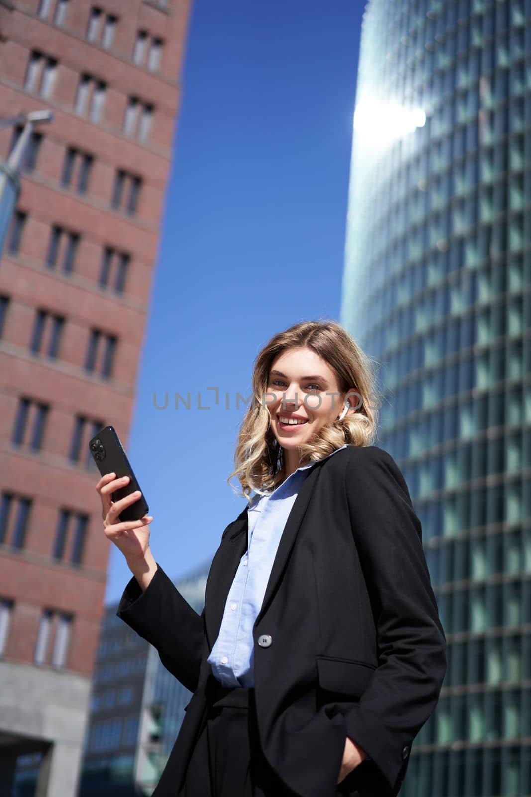 Vertical shot of successful businesswoman in suit, holding smartphone, using mobile phone on her way to work, standing with telephone on street.