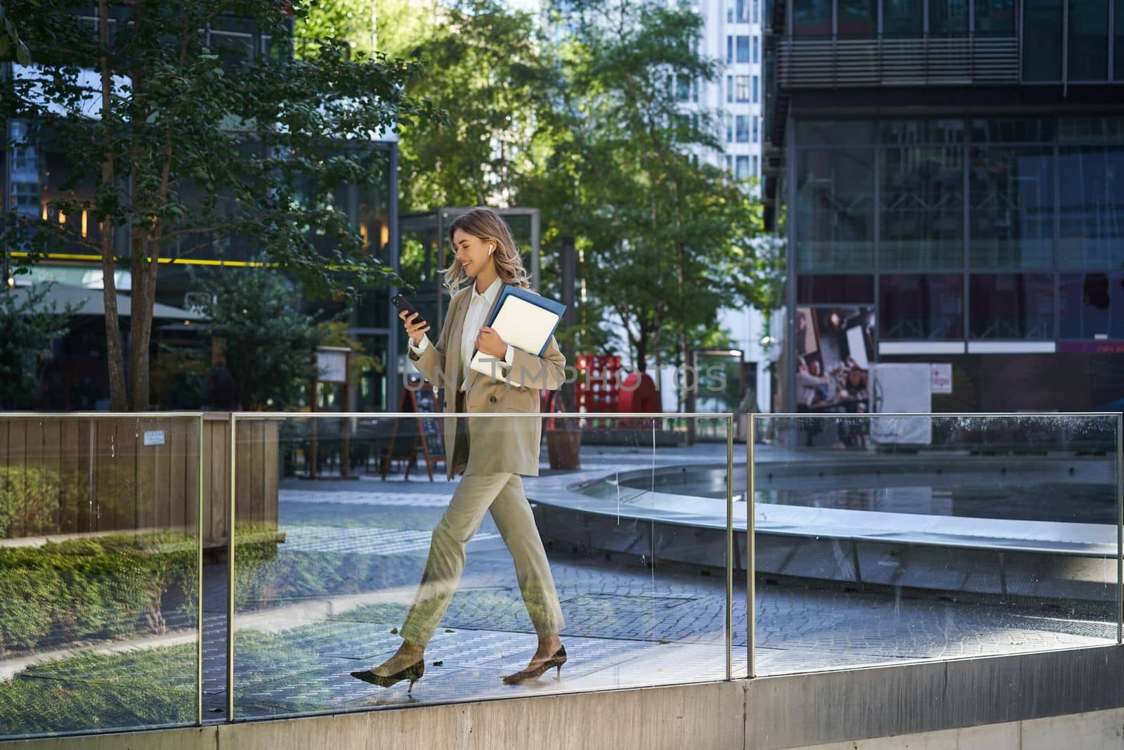 Businesswoman in suit, walking in city, going to work with laptop and documents. Corporate people concept by Benzoix