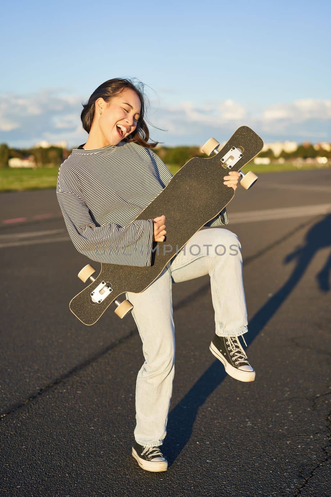 Funny asian girl enjoying skating, holding skateboard like guitar and shadow playing, having fun outdoors by Benzoix