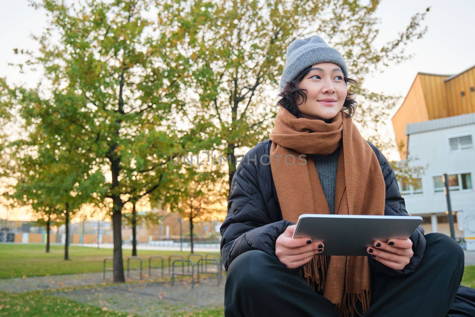 Portrait of asian girl in warm clothes, sits on bench with digital tablet and graphic pen, smiling happily, draws outdoors in chilly weather.