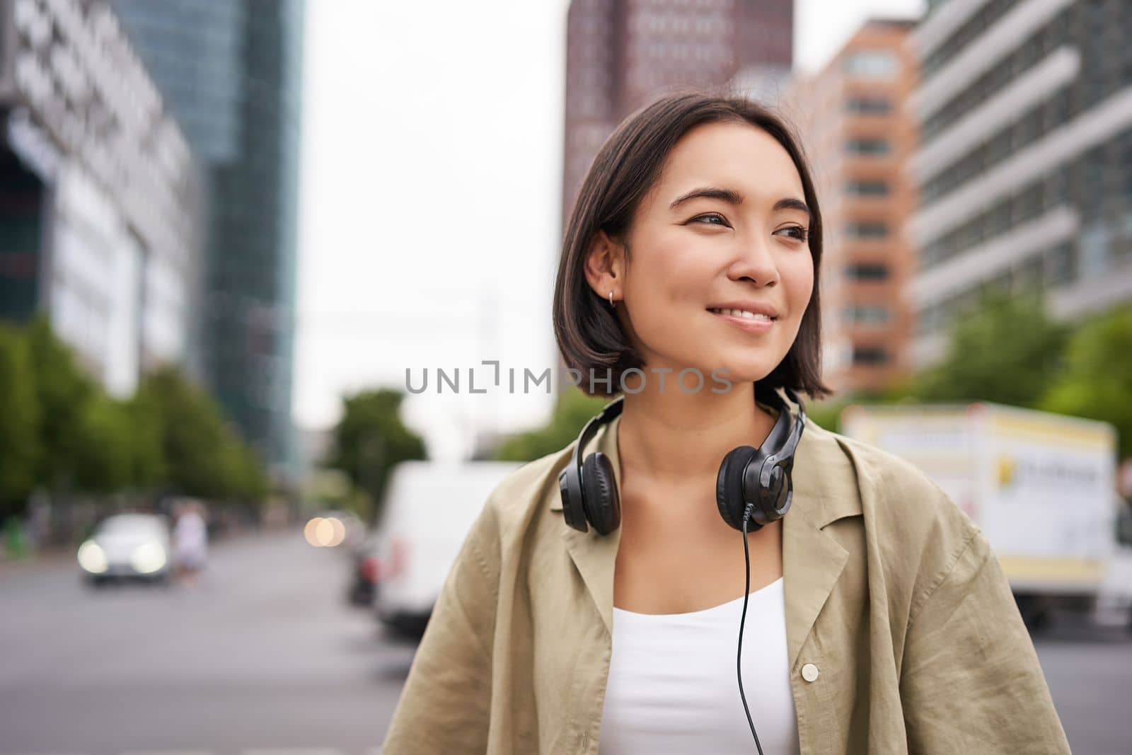 Portrait of young asian woman in headphones, posing in city, smiling and looking away, standing on street of city centre.