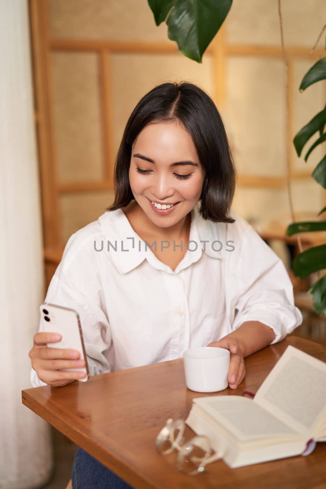 Happy asian woman sits in cafe with cup of coffee and book, answer video call on smartphone, laughing and talking via camera on mobile phone.