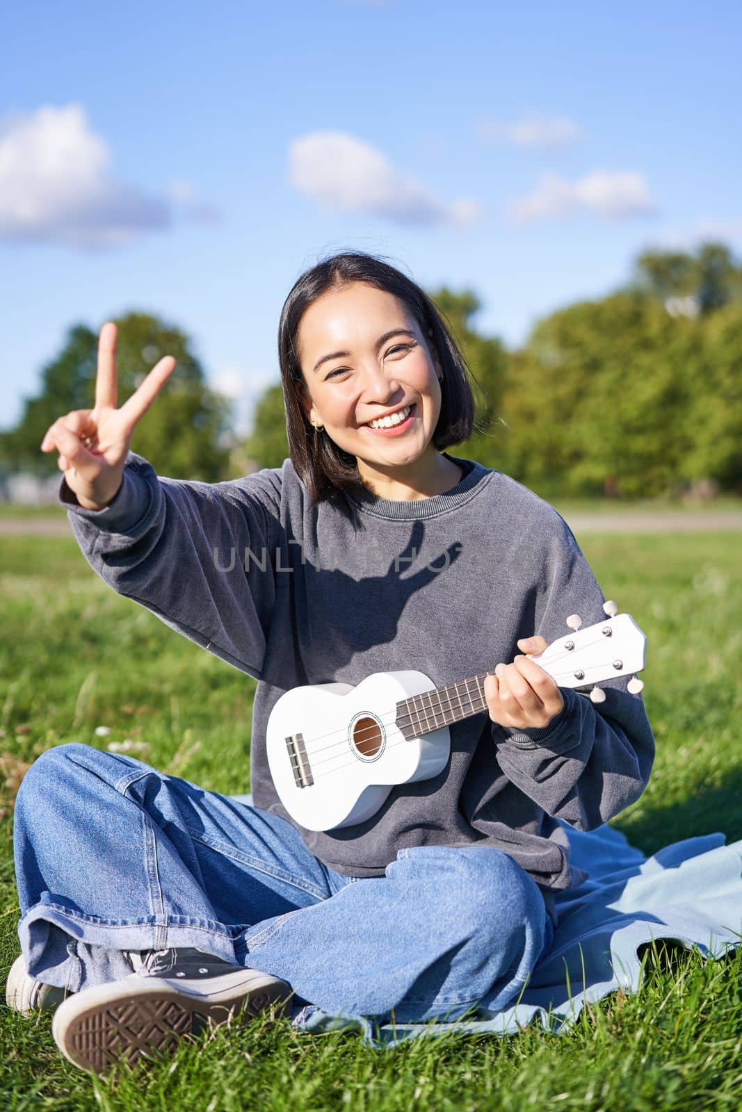 Vertical shot of positive asian girl shows peace sign, plays ukulele in park, rests and enjoys the day. Copy space.