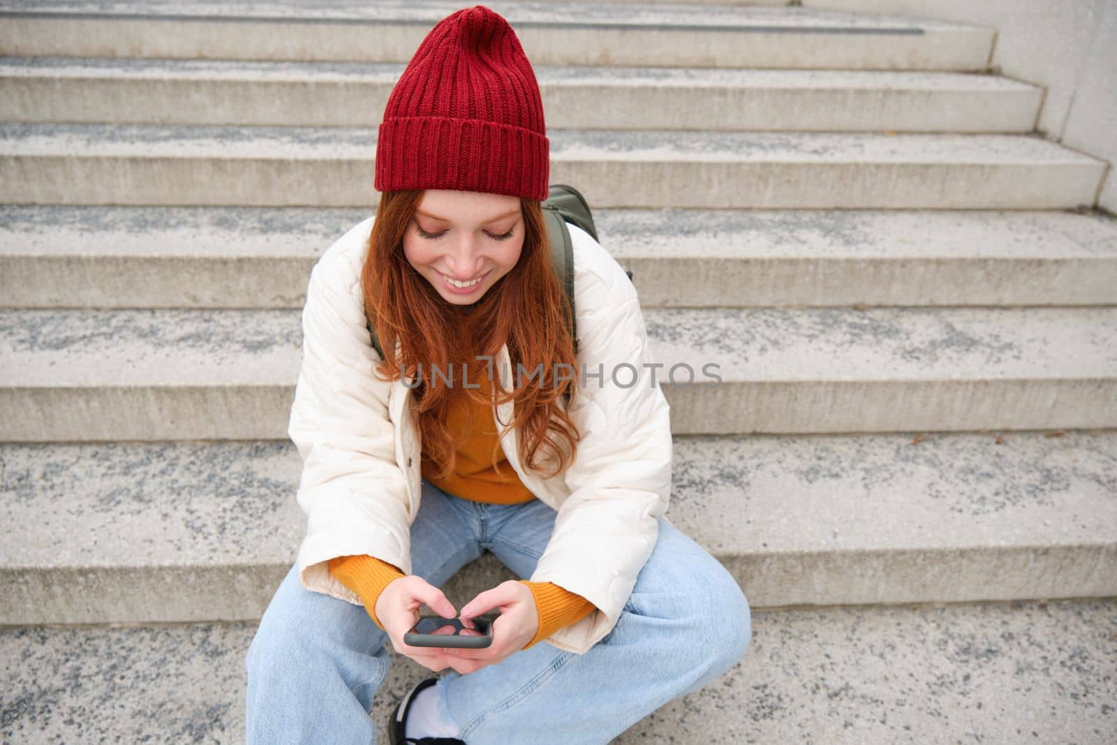 Hipster ginger girl, redhead woman sits on stairs with smartphone, waits for someone and messages on social media on mobile phone app. People and technology concept