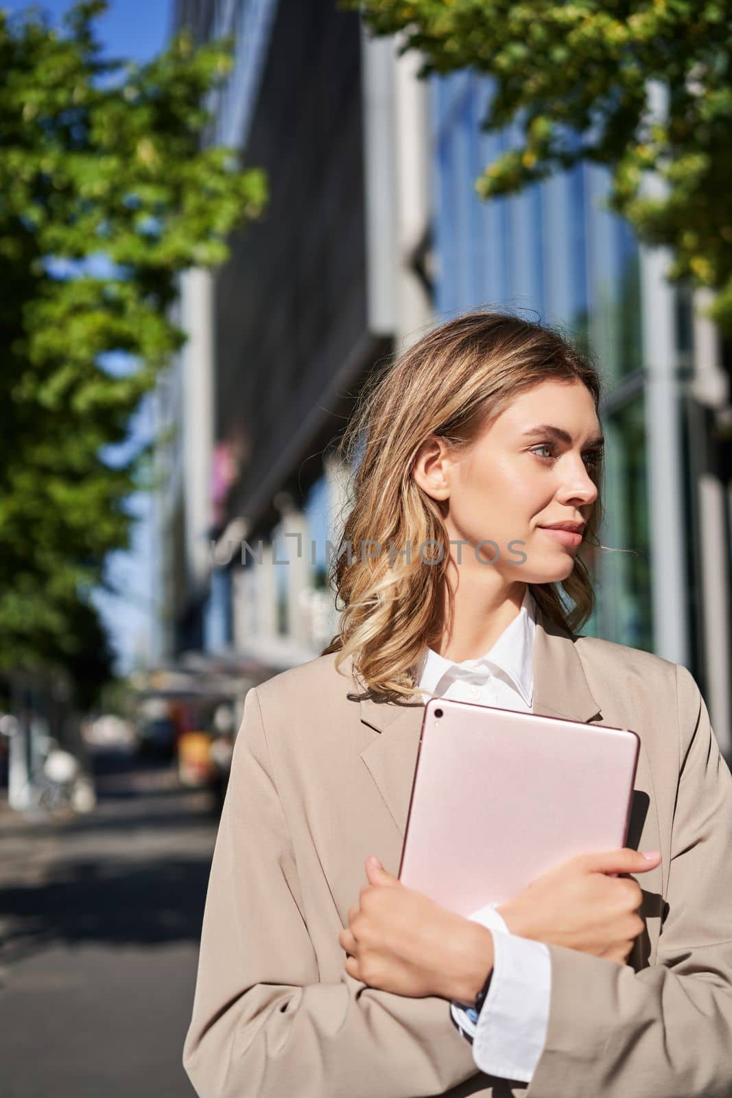 Vertical portrait of corporate woman holding digital tablet, posing on street, adjusting her hair and smiling by Benzoix