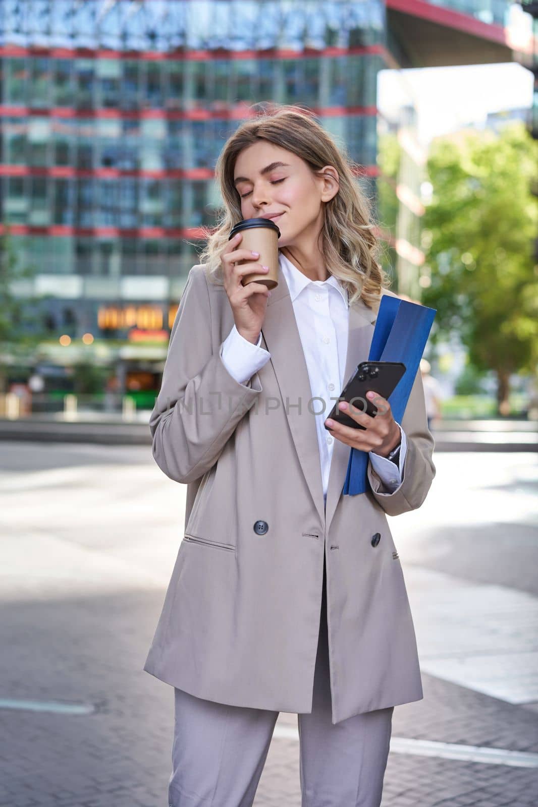 Vertical shot of businesswoman drinking coffee on street, standing in beige suit, holding documents and mobile phone.