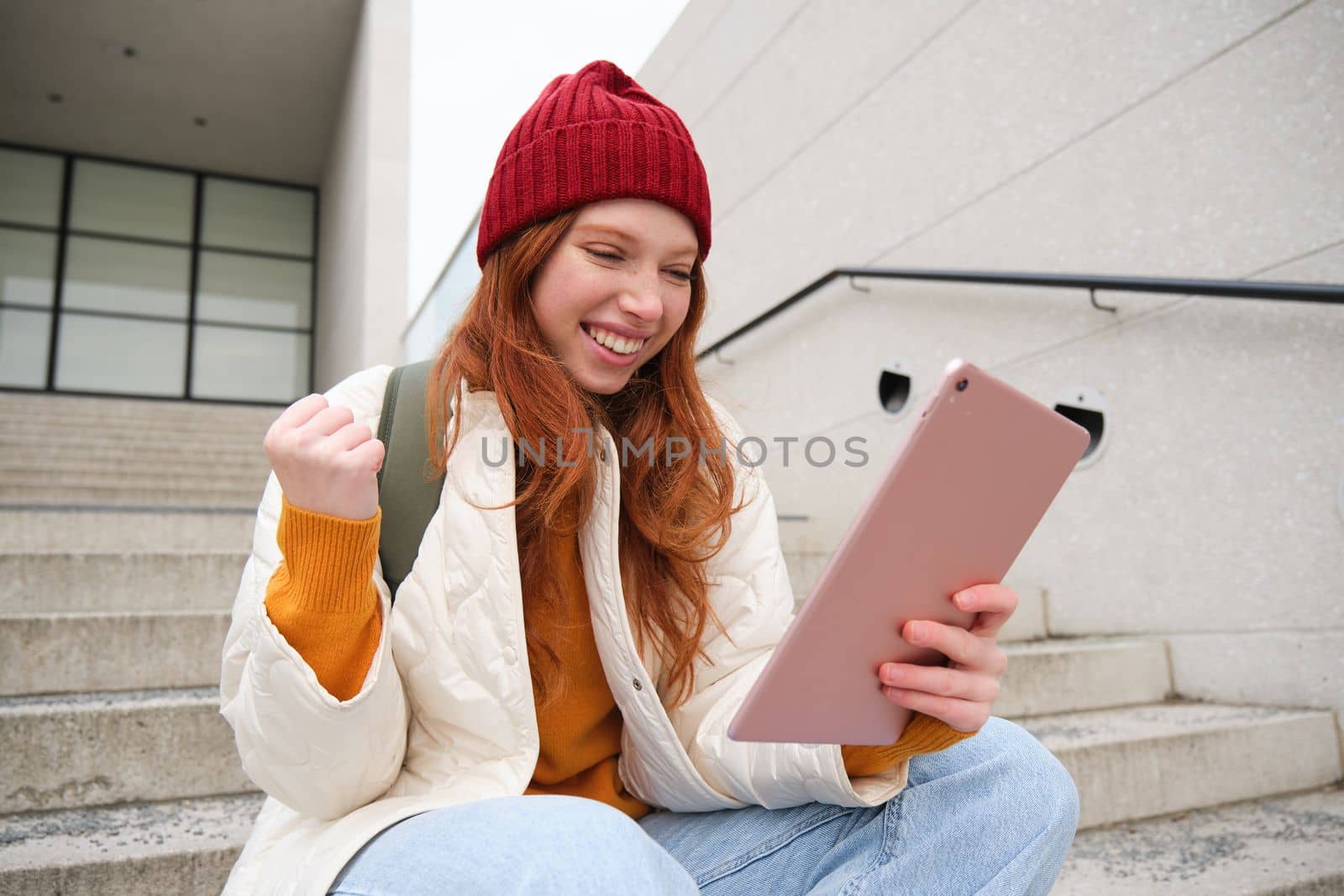 Young stylish girl, redhead female students sits on stairs outdoors with digital tablet, reads, uses social media app on gadget, plays games while waits on street.