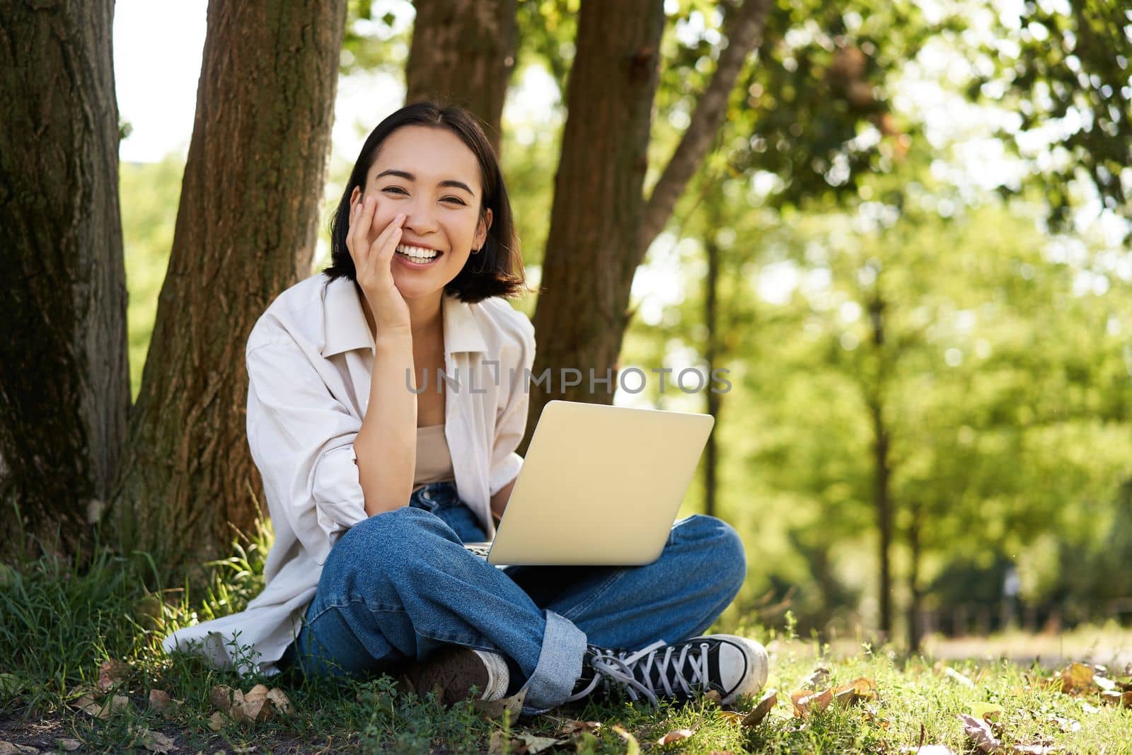 Portrait of young asian woman sitting in park near tree, working on laptop, using computer outdoors.