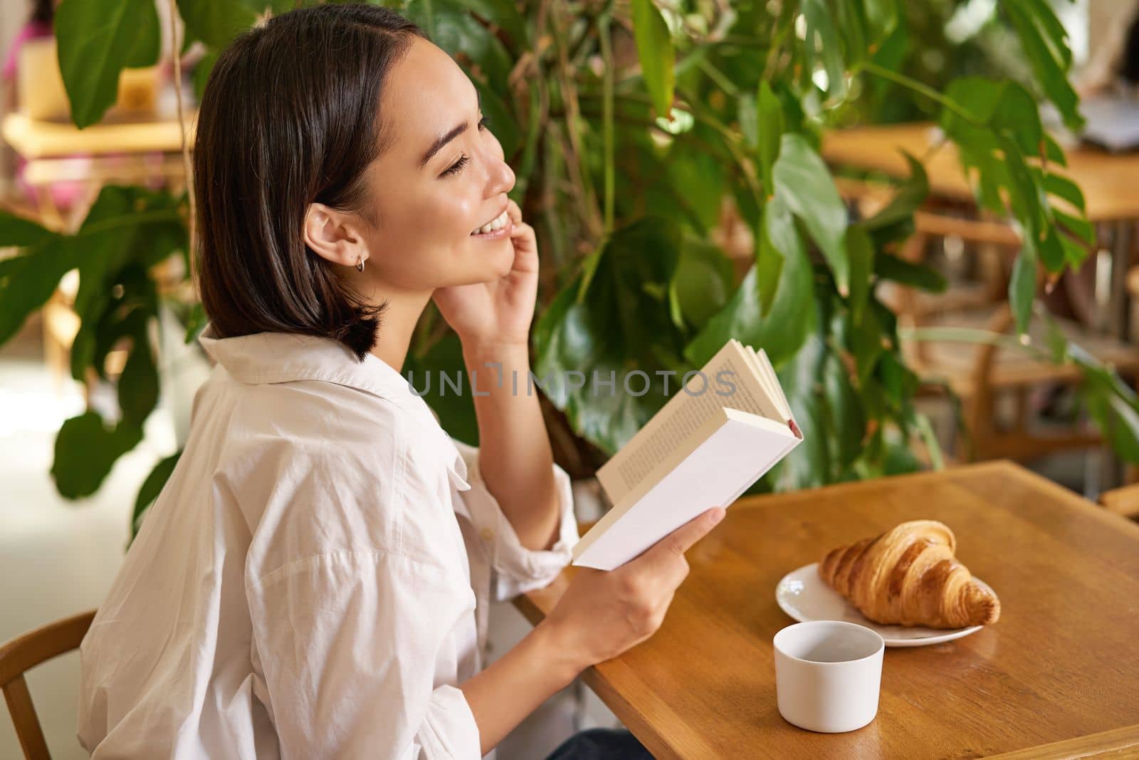 Beautiful young asian woman with a book in hands, sitting in cafe, drinking coffee and eating croissant, smiling, looking mysterious.