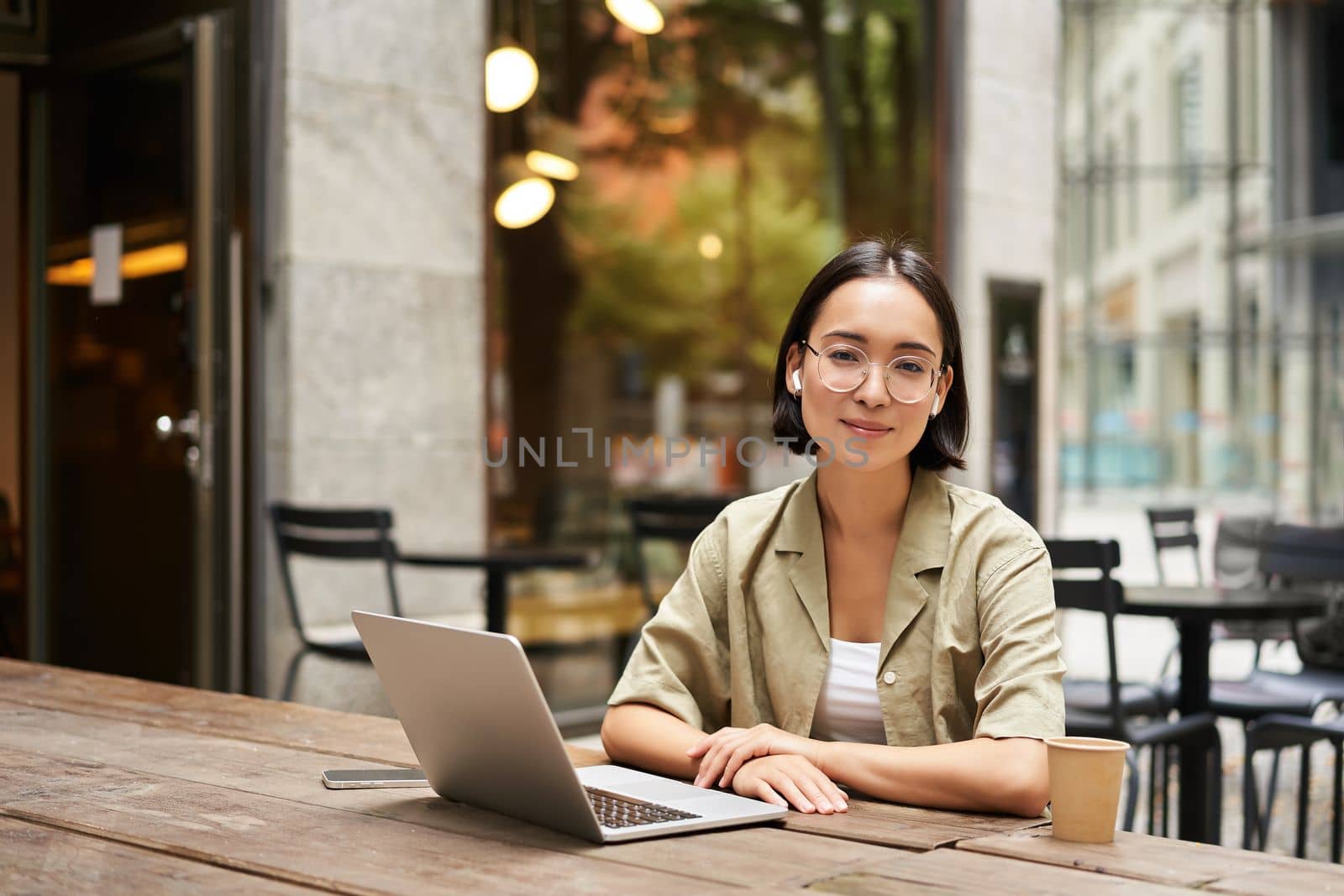 Young woman, student working from cafe, sitting with laptop and cup of coffee outdoors, looking at camera.