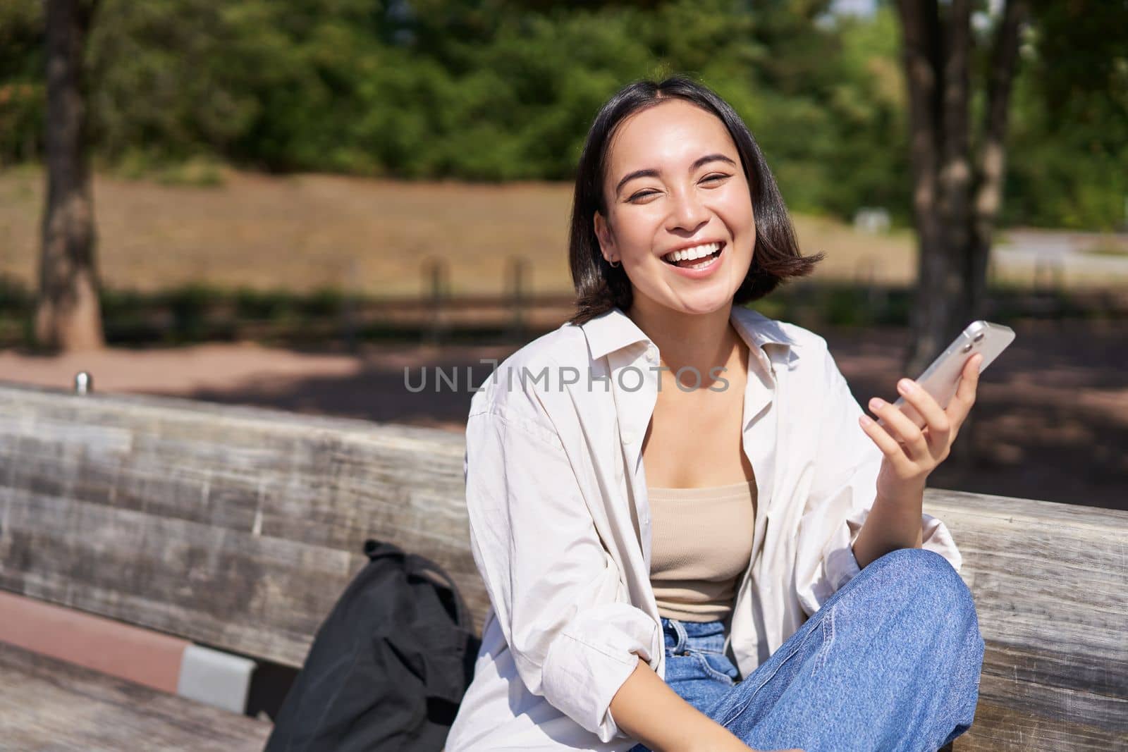 Portrait of happy young asian woman talking on mobile phone in park, sitting on bench and having a telephone call, chatting lively.