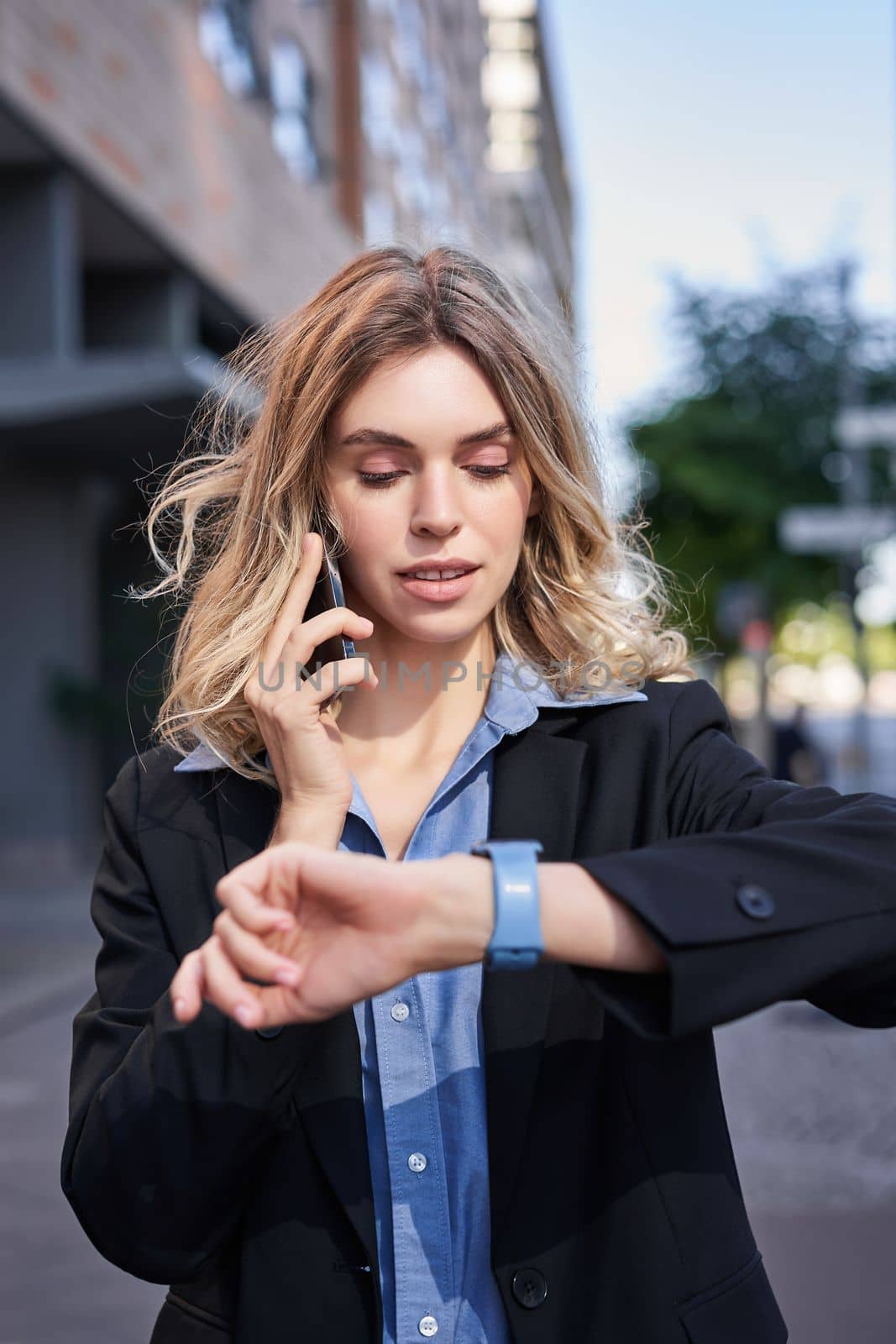 Portrait of confident businesswoman going on a meeting, standing on street, talking on mobile phone and checking time on digital watch.