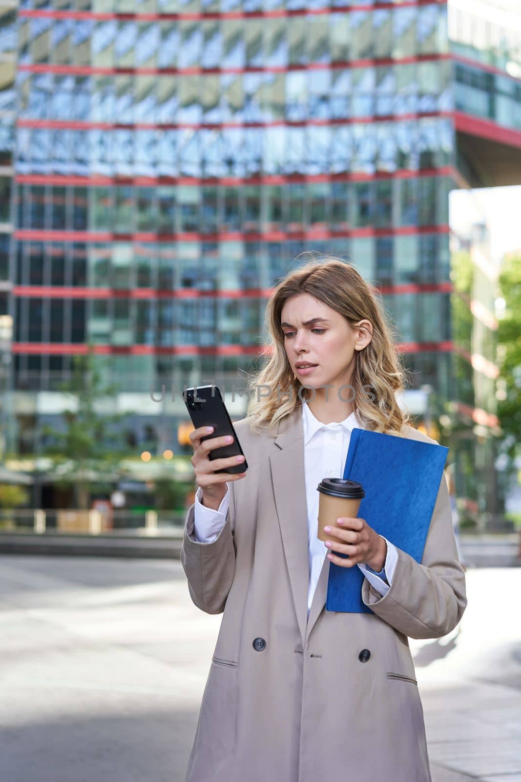 Vertical shot of corporate woman reading message on mobile phone, holding documents and drinking takeaway coffee, standing outdoors by Benzoix