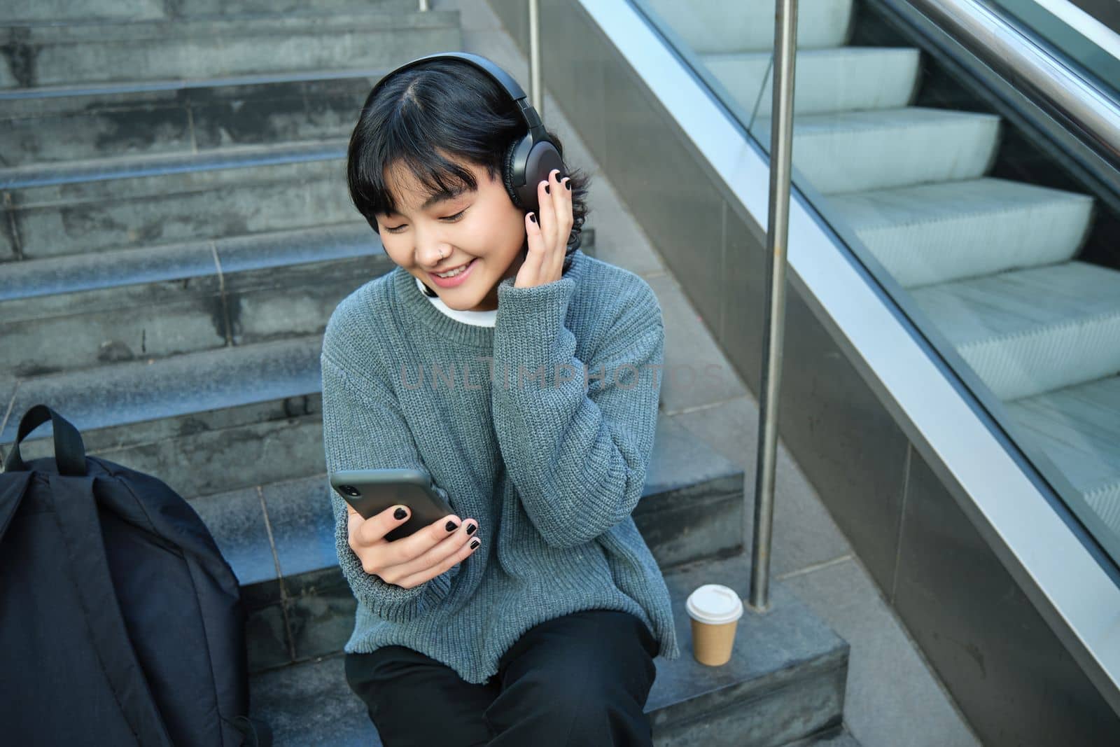 Portrait of smiling korean girl in headphones, uses smartphone and sits on stairs in mall, watches video on mobile phone. Copy space