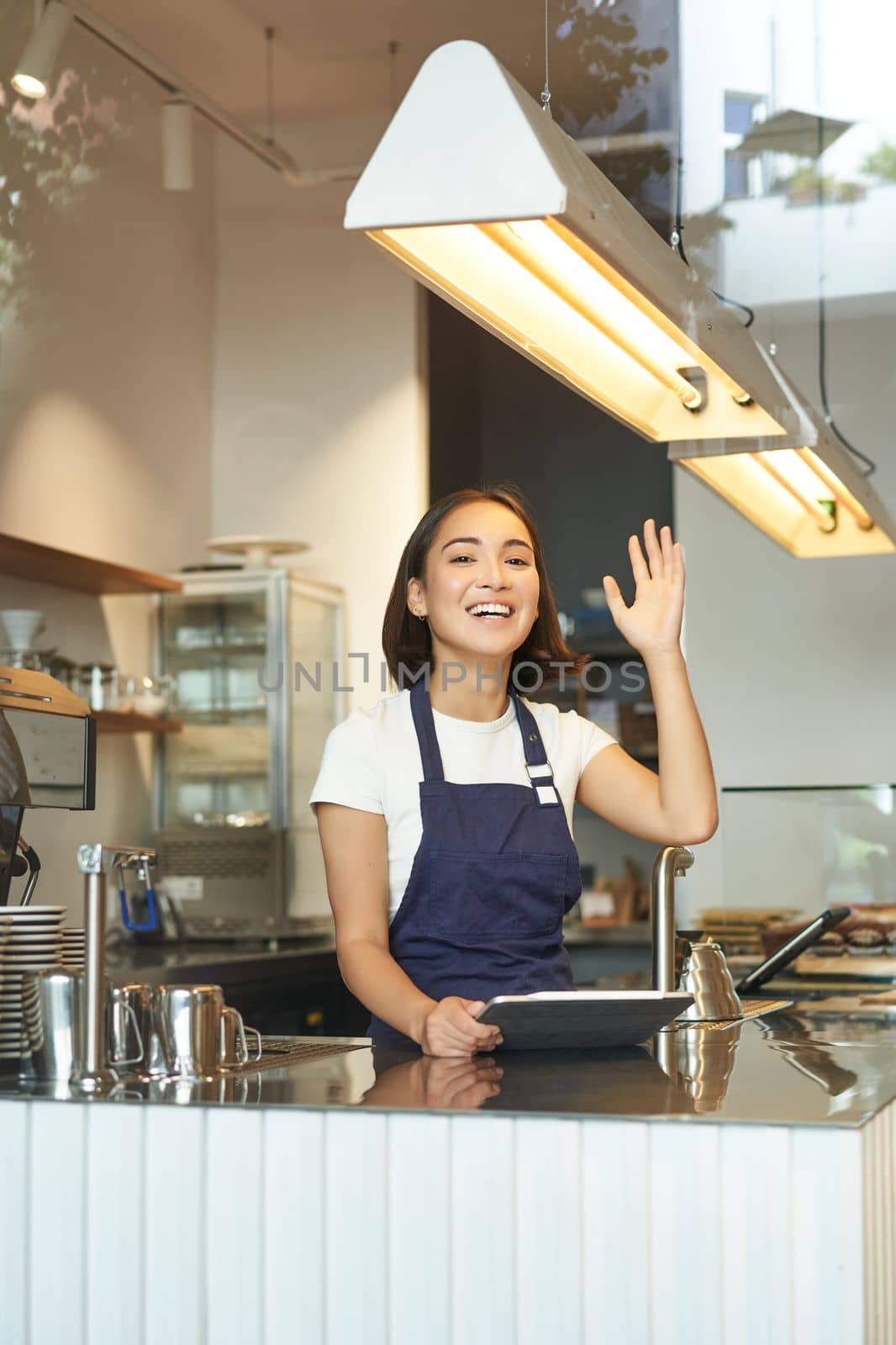 Vertical shot of happy young barista, asian cafe worker waves at client, receives orders behind counter, using tablet as POS terminal, working in coffee shop.